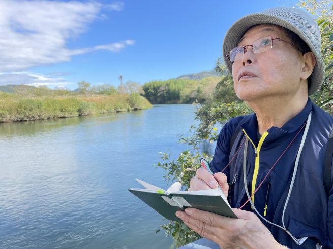 川岸でスケッチをする村松さんのアップ。背景には川